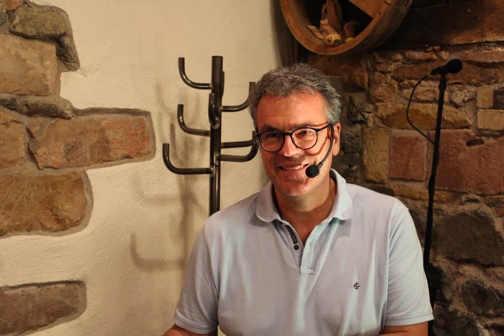 Ein lächelnder Martin Guth mit Brille und Headset-Mikrofon sitzt vor einer rustikalen Steinwand.