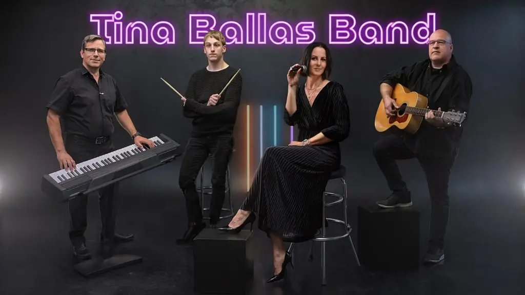 Die Tina Ballas Band posiert mit Sängerin, Keyboard, Schlagzeugsticks und Gitarre vor dunklem Hintergrund.