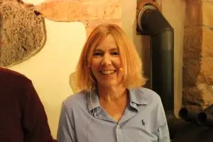 Uta Rosa Schmidt lächelnd, mit Headset-Mikrofon, steht vor einer rustikalen Steinwand.