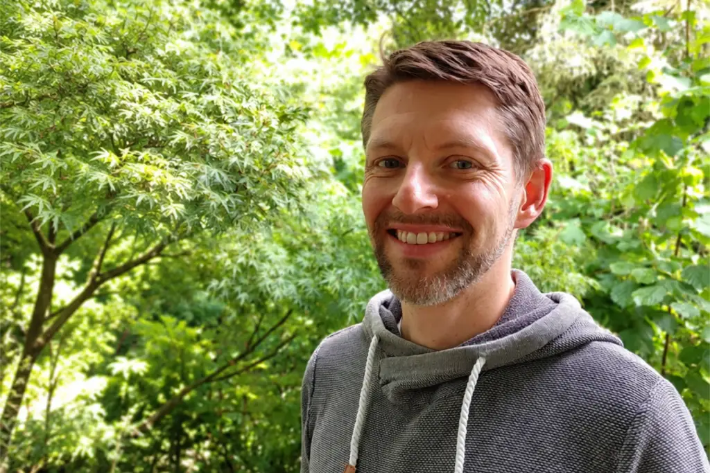 Porträt eines lächelnden Mannes im grauen Pullover vor einer grünen Gartenkulisse.