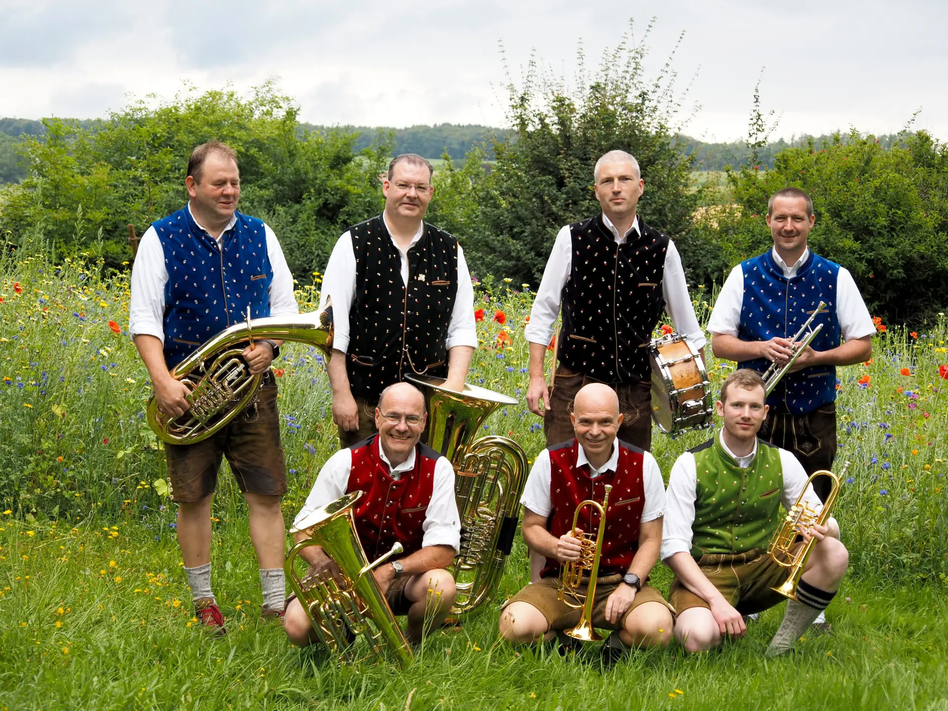 Sieben Musiker in traditioneller Tracht posieren mit verschiedenen Blechblasinstrumenten auf einer Blumenwiese.