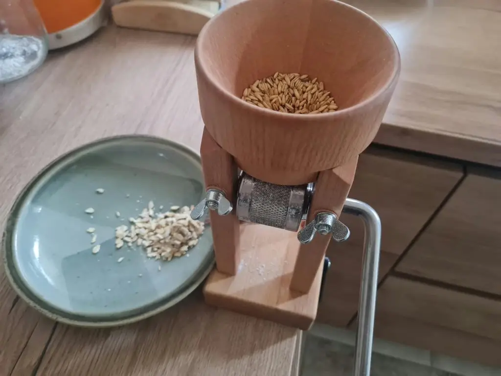 Kleine handbetriebene Getreidemühle mit Körnern im Trichter und frisch gequetschtem Getreide auf einem Teller.
