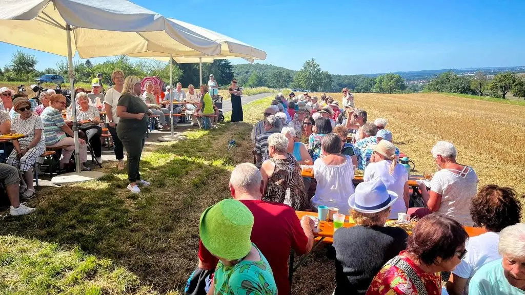 Große Gruppe von Menschen sitzt an Bierzeltgarnituren bei einer Veranstaltung unter Sonnenschirmen am Feldrand.
