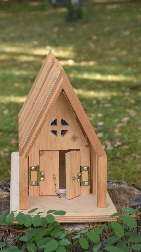 Kleines Holzmodell der Lichtkirche mit spitzem Dach, Kreuzfenster und geöffneten Türen, aufgestellt im Grünen im Kurpark Bad Salzhausen.