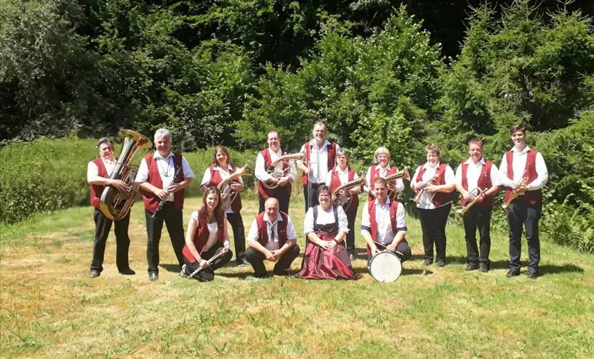 Musikgruppe in rot-weißer Tracht mit Blechblasinstrumenten auf einer Wiese vor grüner Baumkulisse.