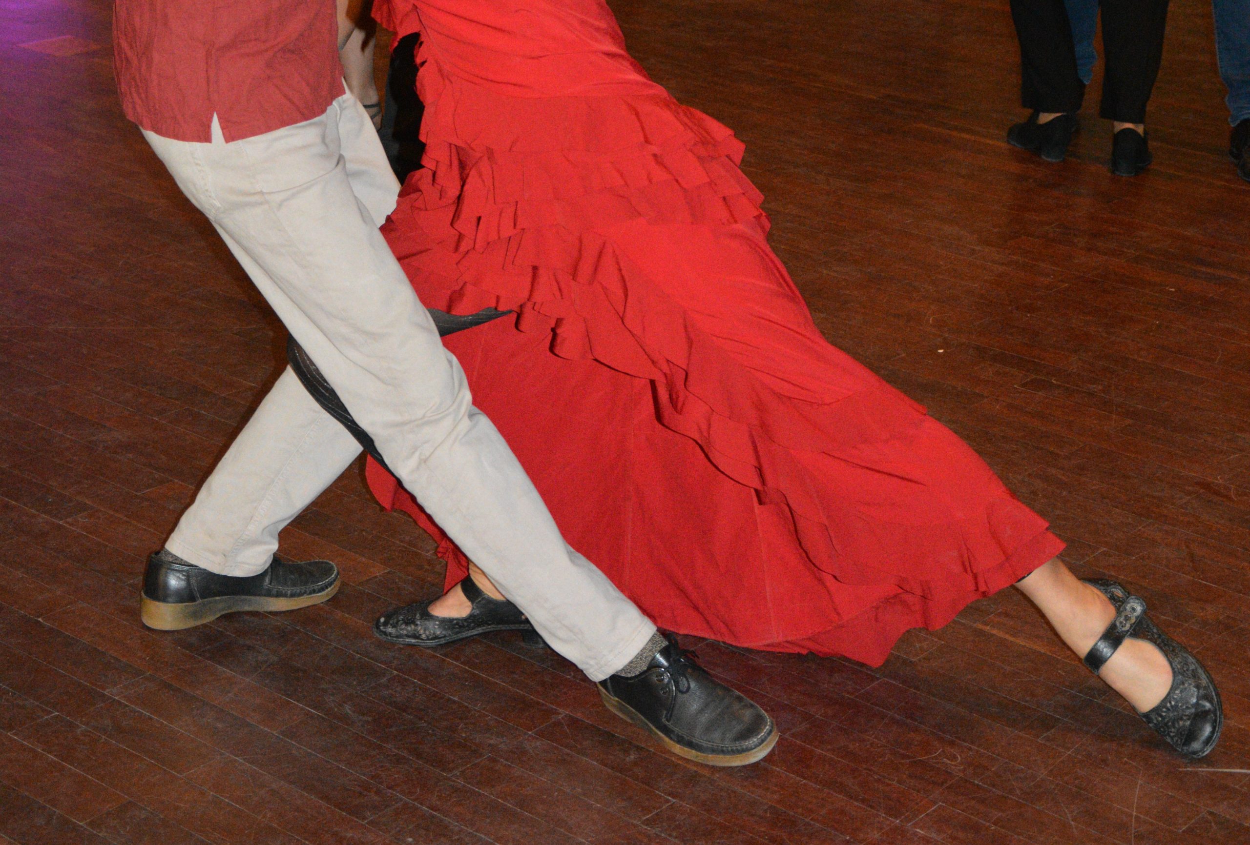 Nahaufnahme eines tanzenden Paares, bei dem vor allem die gekreuzten Beine und das rote Kleid zu sehen sind.