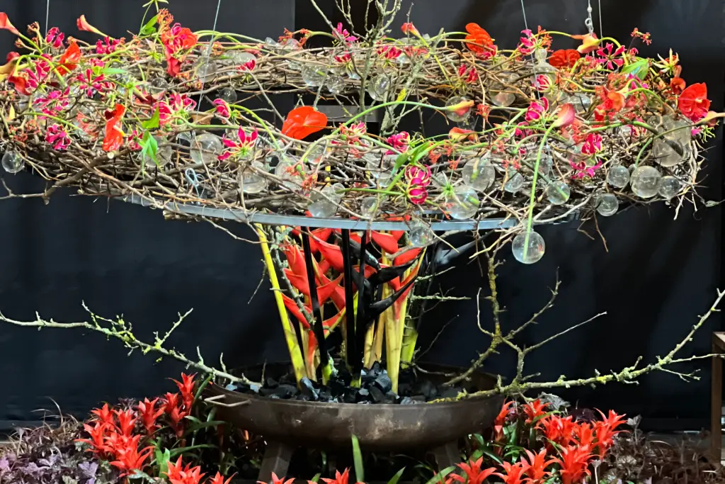 Aufwendiges florales Arrangement aus Zweigen, roten Blüten und Glaskugeln vor dunklem Hintergrund.