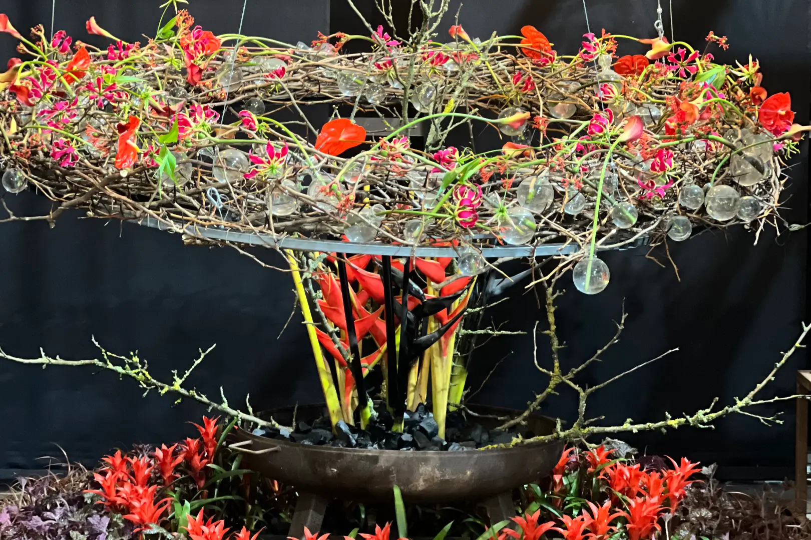 Aufwendiges florales Arrangement aus Zweigen, roten Blüten und Glaskugeln vor dunklem Hintergrund.